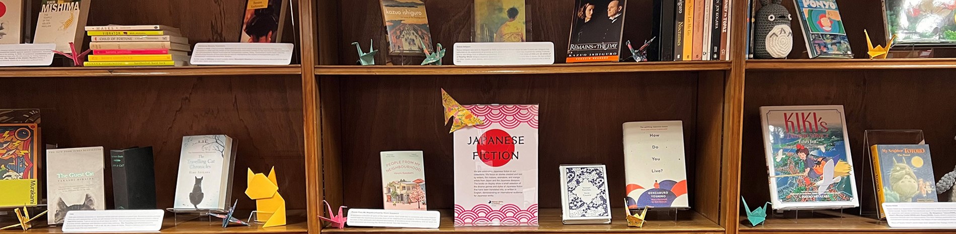 A photo of a bookshelf filled with Japanese books. There are also several origami paper cranes scattered on the shelf near the books. There is a sign in the middle saying "Japanese fiction".