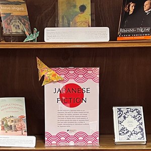 A photo of a bookshelf filled with Japanese books. There are also several origami paper cranes scattered on the shelf near the books. There is a sign in the middle saying "Japanese fiction".