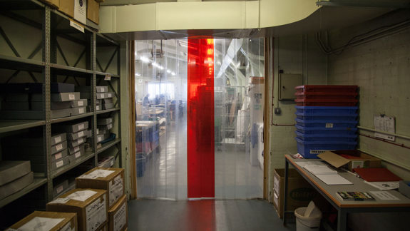 Storage room with metal shelves filled with boxes and files on the left, cardboard boxes on the floor, a transparent plastic strip curtain with a red center strip in the middle, and a table with papers and plastic trays on the right.
