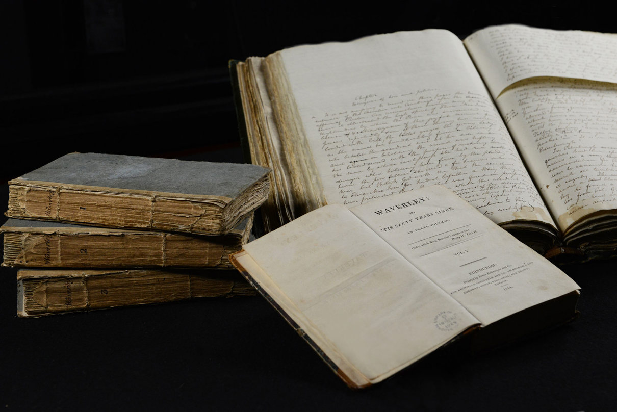 A display of old books and manuscripts. On the left, four closed books are stacked. In the center is an open copy of Walter Scott’s Waverley, and to the right are two large open manuscripts with handwritten text.