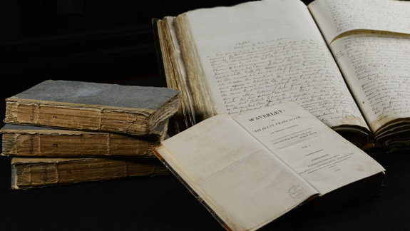 A display of old books and manuscripts. On the left, four closed books are stacked. In the center is an open copy of Walter Scott’s Waverley, and to the right are two large open manuscripts with handwritten text.