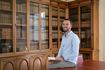 A man holding a book sitting on a table in a room full of books.