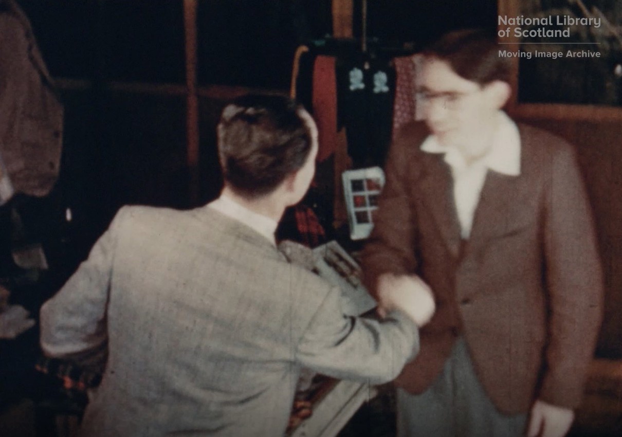 Two men shake hands in an indoor setting with various tartan items and clothing in the background. One man faces away from the camera; the other, faces toward it.