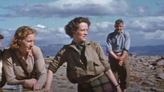 Three young women in the foreground and a man in the background on the top of a mountain with hills behind them.