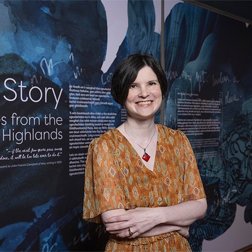 Photo of Kirsty MacDonald standing in front of a panel for an exhibition which reads "Sgeul | Story. Folktales from the Scottish Highlands".