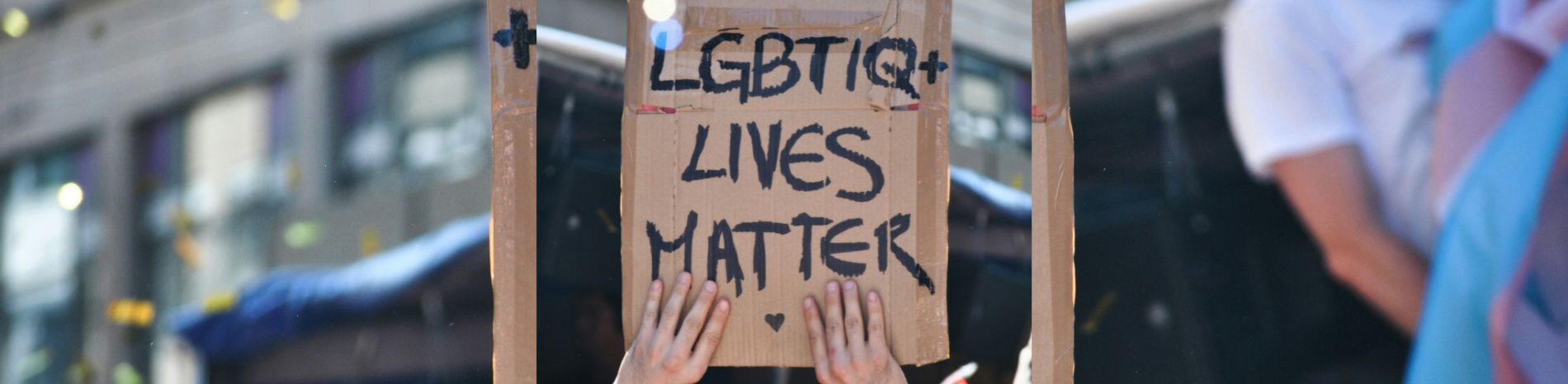 A homemade cardboard placard saying "LGBTIQ+ lives matter". There is a blurry trans flag in the background on the right.