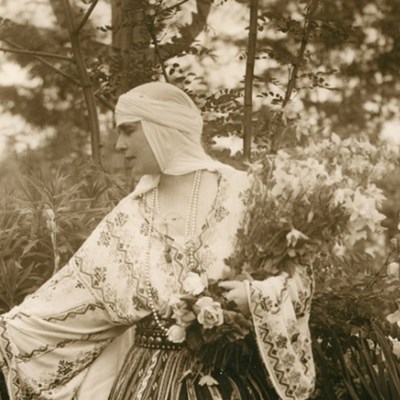 Princess Marie stands in a flowing gown in a garden full of flowers. She holds a basket over her arm.