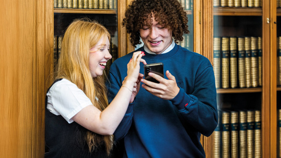 Two students standing in front of a bookcase looking at something on a phone.