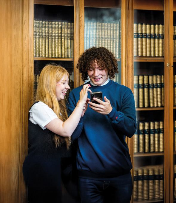 Two students standing in front of a bookcase looking at something on a phone.