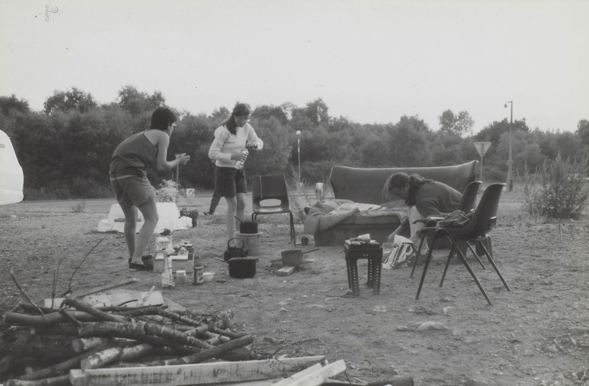 Three people around a campfire surrounded by trees and bushes. One person pours water from a bottle, another bends near a couch, and a third stands with hands clasped.