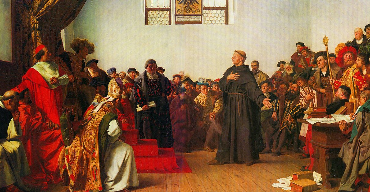 A historical painting showing a large group of people in elaborate, colorful robes gathered in a formal setting. A central monk figure in dark robes stands with one hand on their chest and the other extended outward. The background features two stained glass windows and various individuals seated or standing around the central figure.