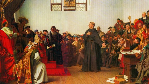 A historical painting showing a large group of people in elaborate, colorful robes gathered in a formal setting. A central monk figure in dark robes stands with one hand on their chest and the other extended outward. The background features two stained glass windows and various individuals seated or standing around the central figure.