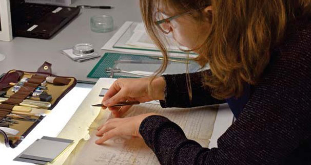 A woman is handling an old manuscript. It is sitting on a lightbox. There is a set of tools in front of her and to the site.