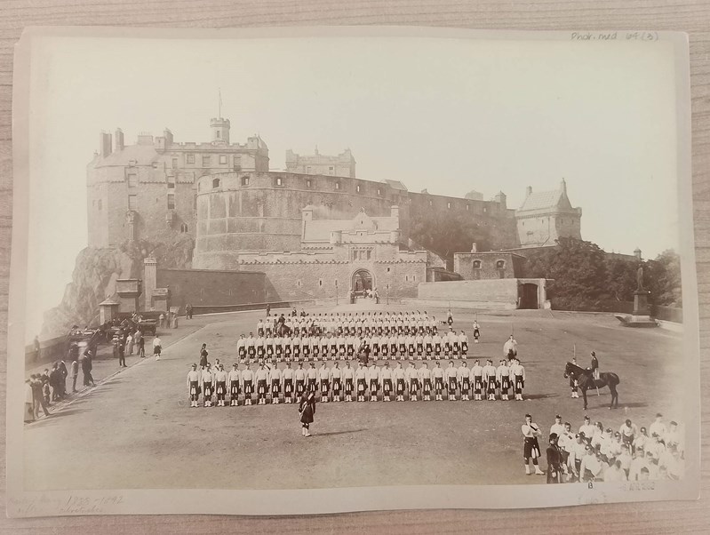 A historical black-and-white photograph showing rows of uniformed soldiers standing in formation on a large open ground. Behind them rises Edinburgh castle with tall walls and towers. A few individuals on horseback and others walking along the edges add to the sense of a formal military gathering.