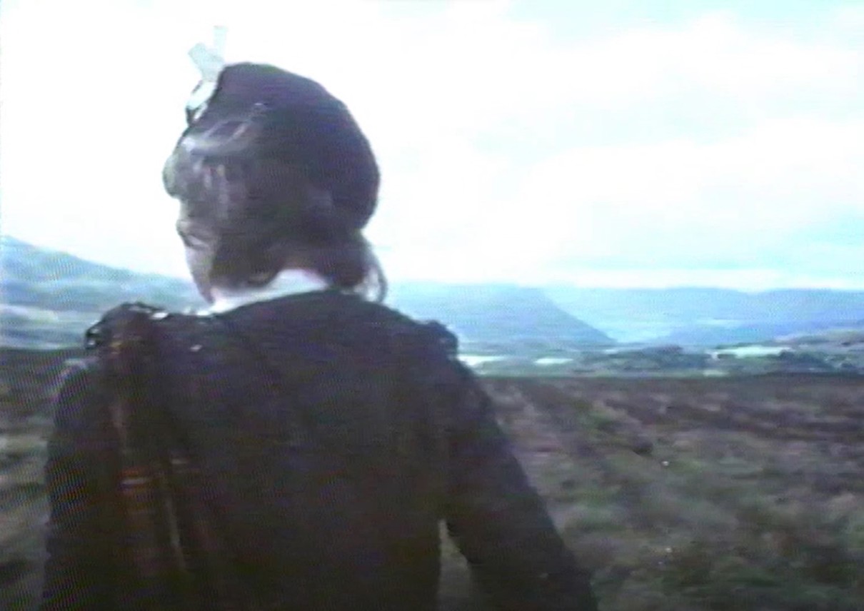Still of a close-up of a person in Highland wear with their back to the camera and moorland beyond.