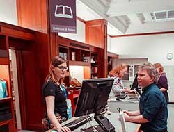 Someone standing behind a desk with a computer while another person watches