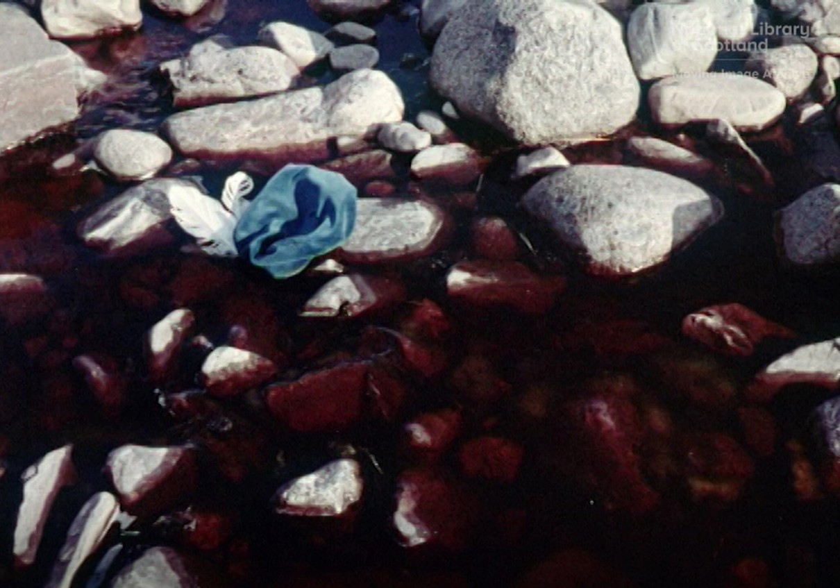 Still showing a close-up of rocks in a river that is running red, with a blue hat with a white feather on it slumped on one of the rocks.
