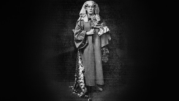 Black and white photo of a man standing in occult garb, including a head piece and a leopard pelt.