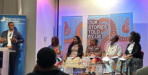 A presenter standing to the left. There are five women sitting down as part of a panel. In the background is a banner which says "Our stories told by us". There is an audience in the foreground.
