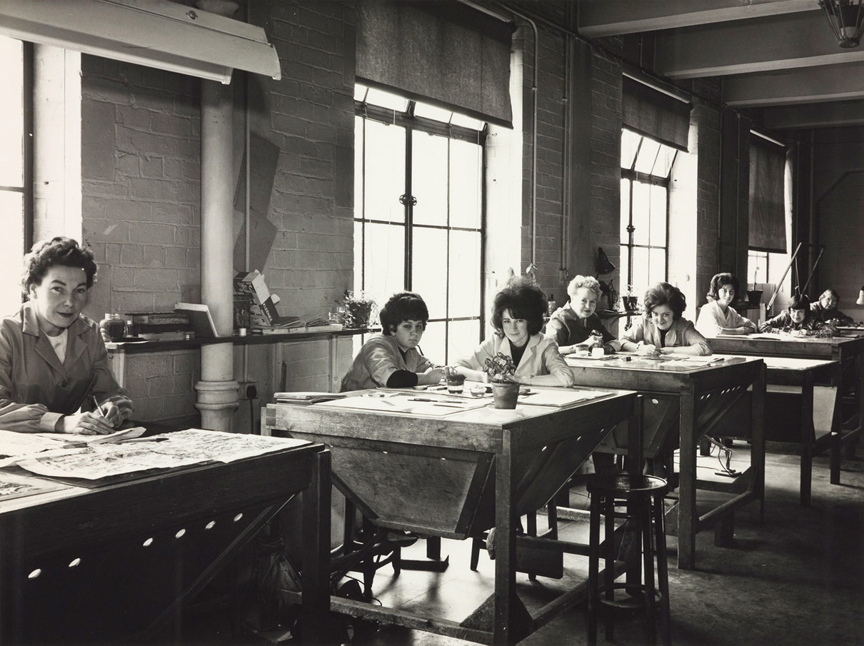 A black and white photo of several women sitting at tables colouring maps by hand.