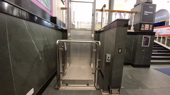 An accessible lift with operating buttons on the right-hand side and a glass door with metal railings to open.