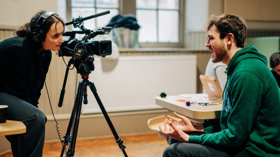 A woman behind a video camera and a man sitting speaking to the camera.