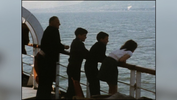 A still from 'In the Lion's Wake' of three children and a man leaning on railings on a ferry, looking out to sea.