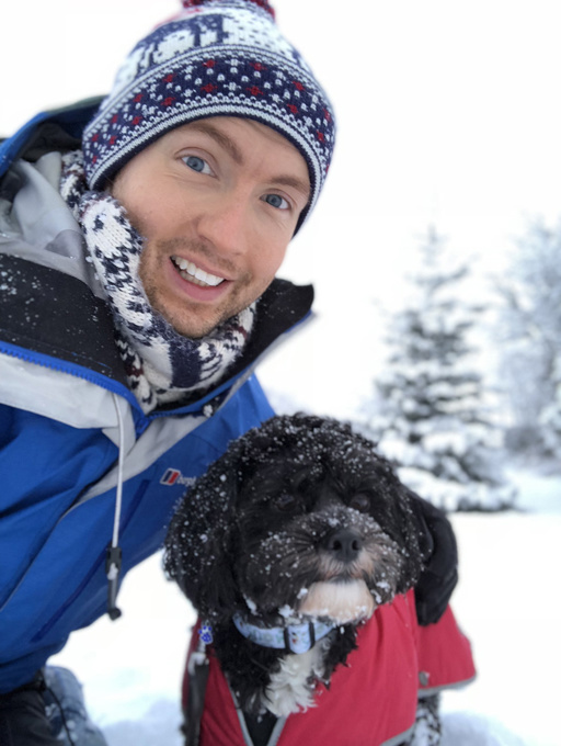 Person in winter clothing posing with a black dog in a red coat in a snowy landscape.