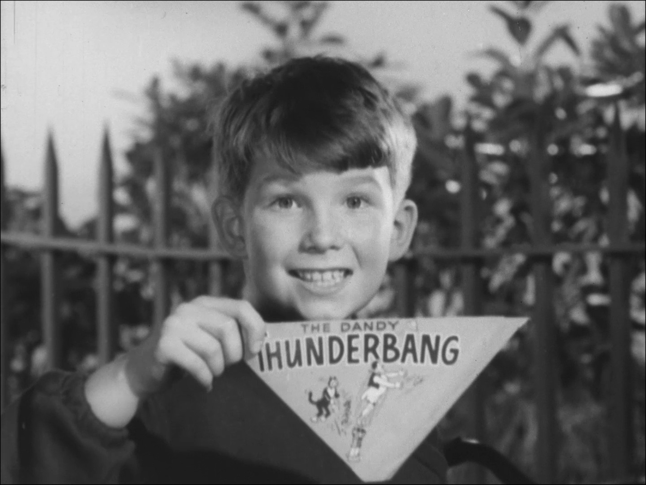 Black and white still from an ad of a little boy smiling and holding a triangular piece of card with the word "Thunderbang" written on it.