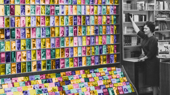 A black and white photo of a woman next to a large shelf of books which have been coloured.