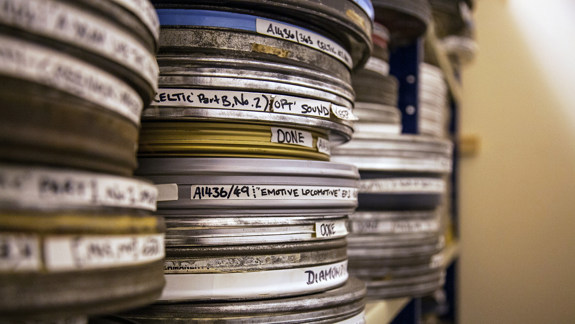 A close up of film reels in metal cannisters with sticker labels on the sides, sitting on a shelf.