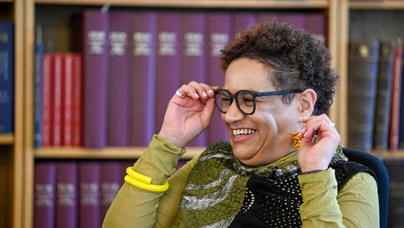 A woman with black glasses smiling and laughing against library shelves. Her hands are raised to touch her glasses.