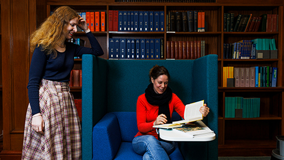 Two library visitors reviewing documents in a reading area with bookshelves behind them.