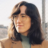 A headshot of a person with shoulder length dark hair, wearing glasses, looking away from the camera.