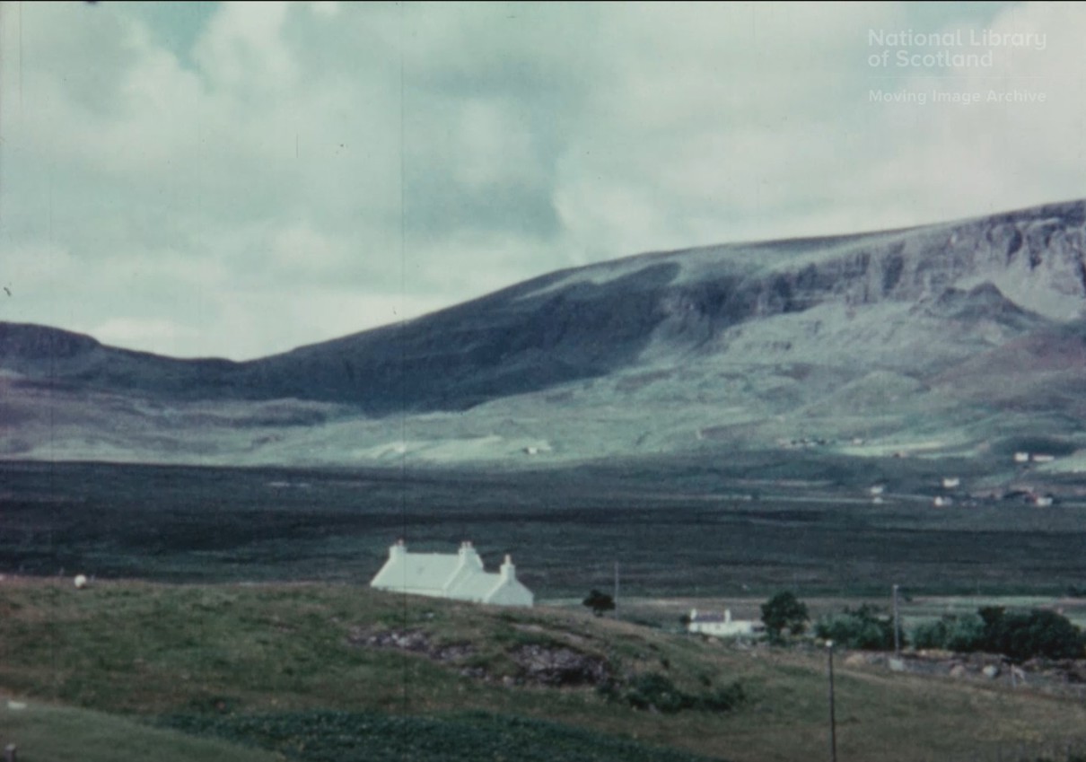 A rural landscape featuring a white house on a grassy hill, with expansive moorland and large hills in the background under a cloudy sky.