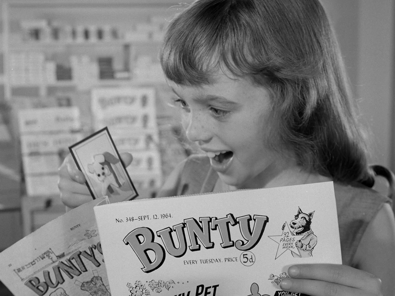 Black and white still from an ad of a little girl holding an issue of Bunty and the free brooch with a puppy on it.