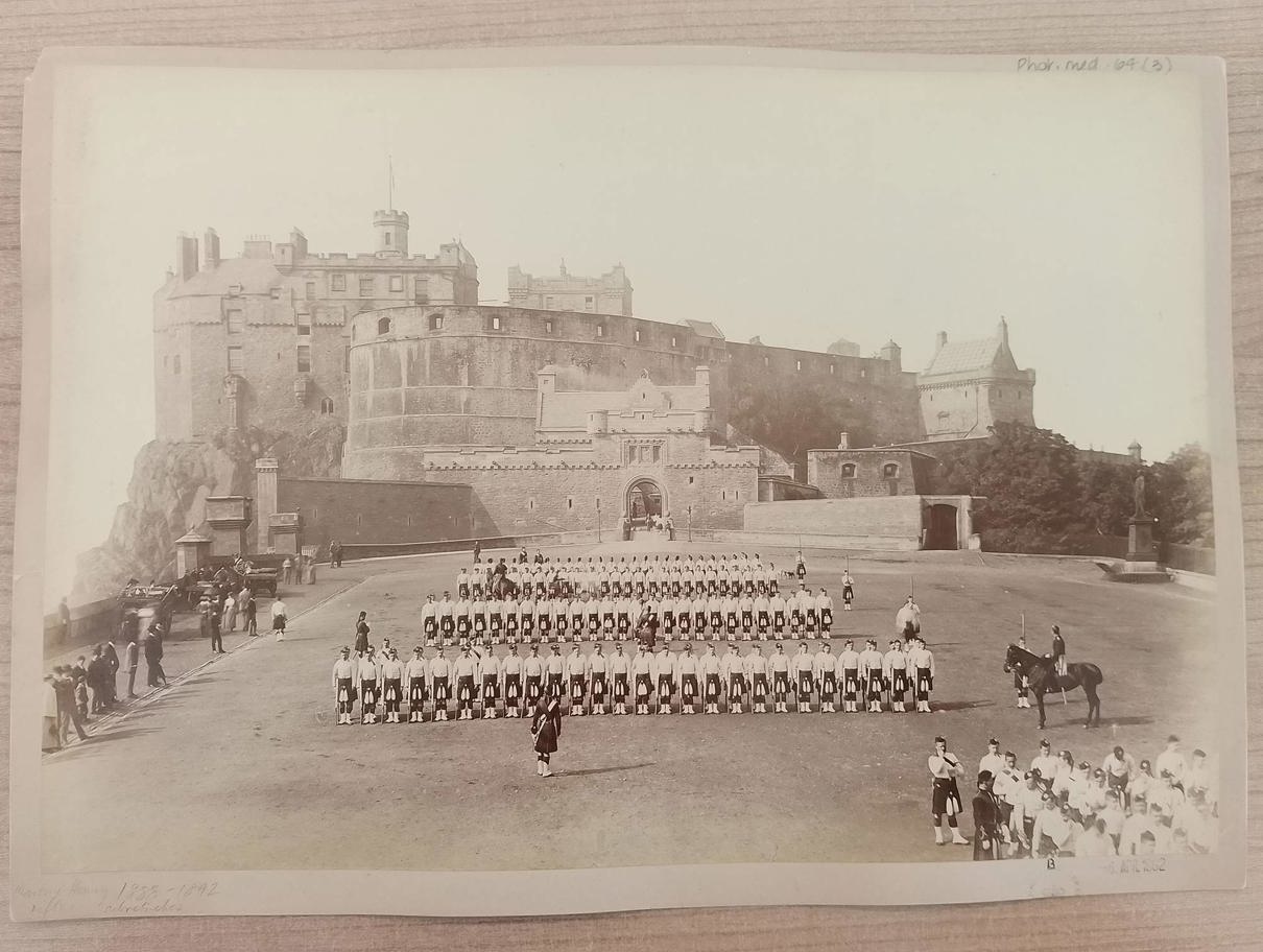 A historical black-and-white photograph showing rows of uniformed soldiers standing in formation on a large open ground. Behind them rises Edinburgh castle with tall walls and towers. A few individuals on horseback and others walking along the edges add to the sense of a formal military gathering.