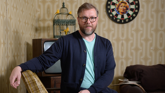 Person seated in a retro-styled room, old TV behind them topped with a birdcage holding two yellow birds, and a dartboard on the wall.