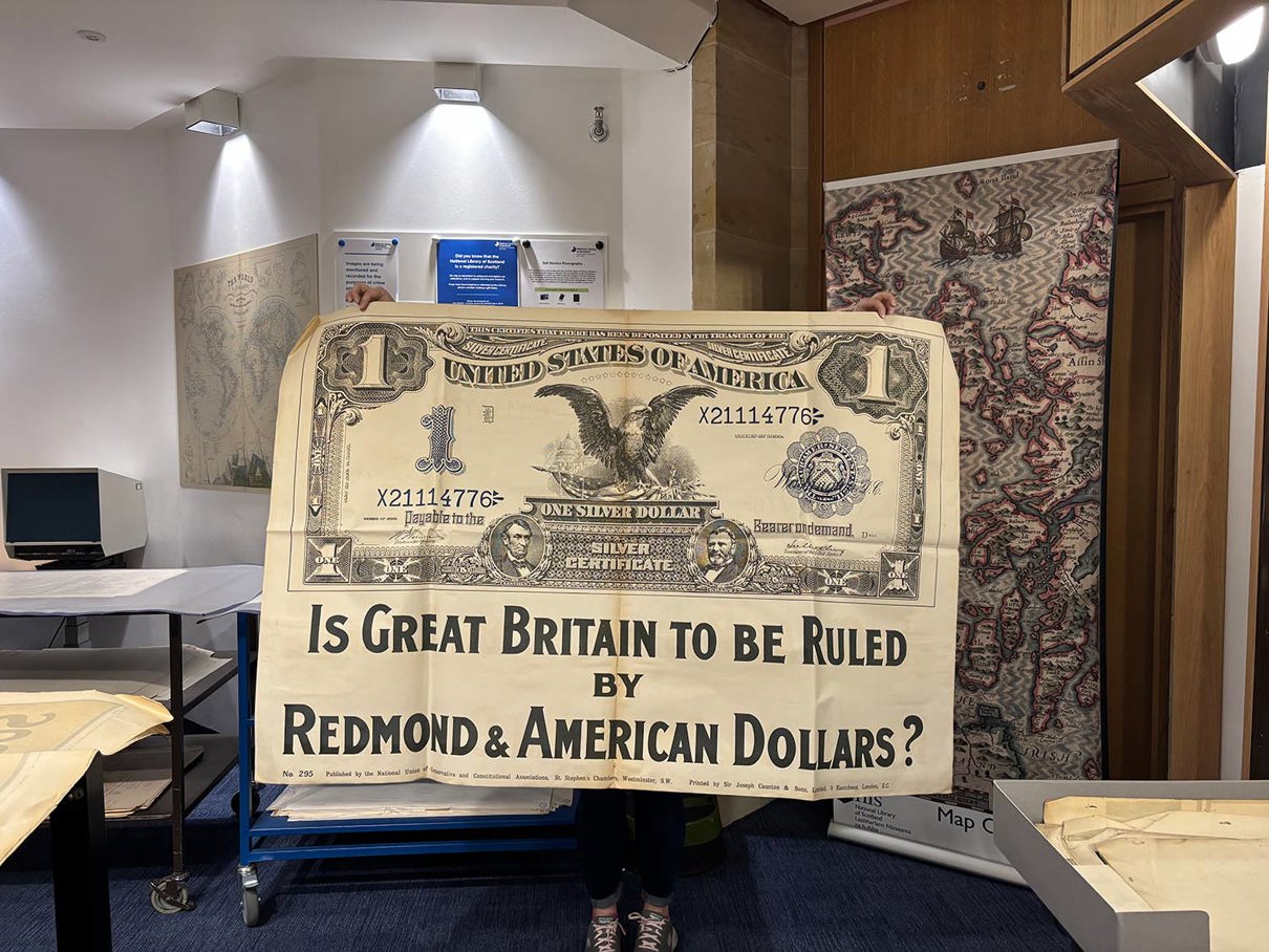 A person holds a large poster designed to look like a vintage U.S. one-dollar bill. It reads: “IS GREAT BRITAIN TO BE RULED BY REDMOND & AMERICAN DOLLARS?” along with typical dollar bill elements like “ONE SILVER DOLLAR” and a serial number.