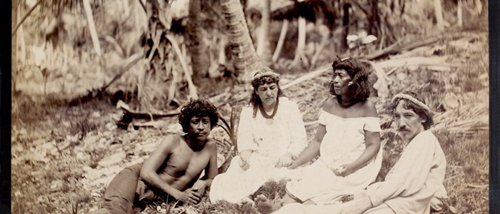A black and white photos of four people sitting on the ground outside. They are surrounded by palm trees.