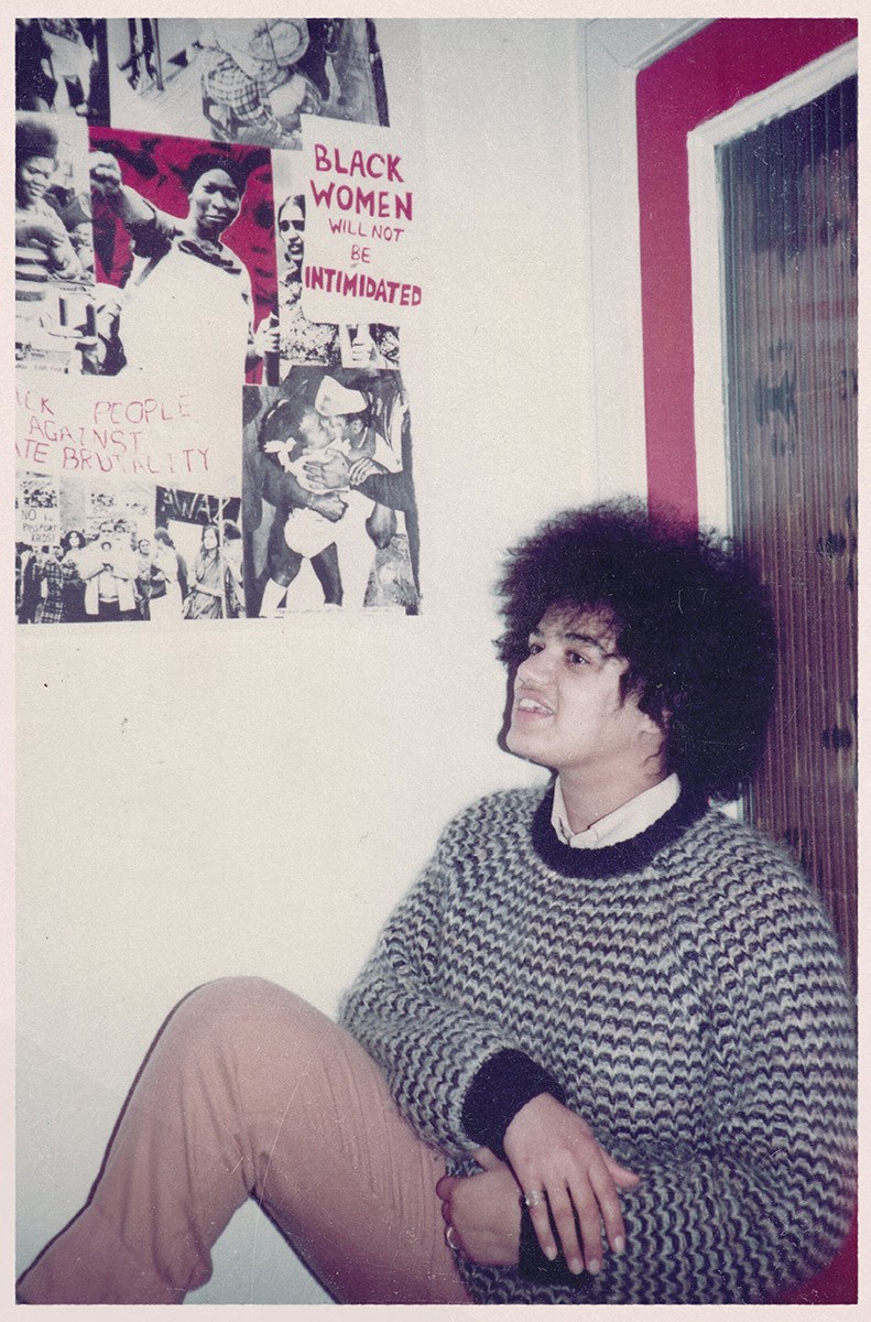 Vintage photograph of Jackie in a striped woolly jumper sitting in the corner against a wall and door. Above her is a collage of black and white imagery alongside handwritten posters, one of which says "Black women will not be intimidated".