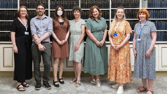 Seven adults standing in a row in a room full of books facing the camera.