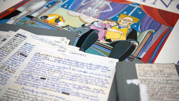 The spread out pages of the handwritten manuscript of 'Poor Creatures' (original name for 'Poor Things'). Underneath these pages is a colourful illustration showing a large man sitting on a couch and and boy and a girl sitting on his lap.