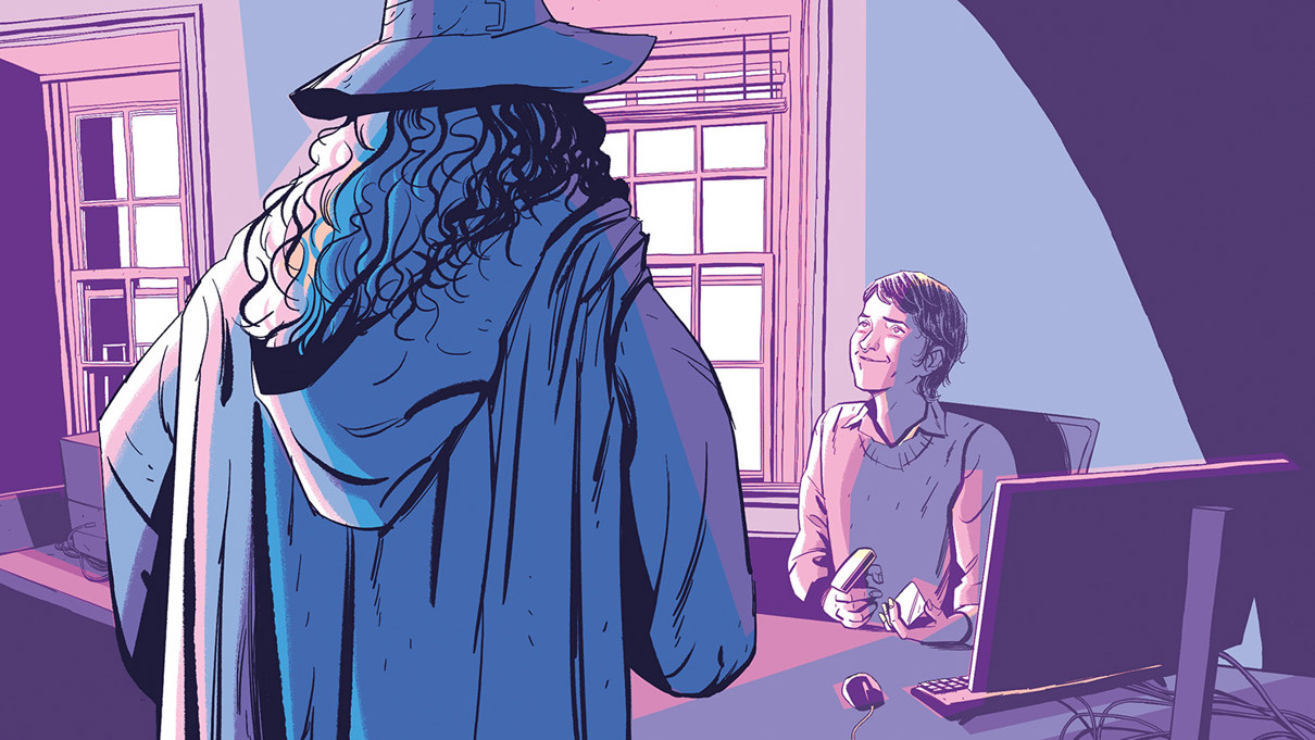 Merlin stands in front of a desk where a member of staff greets him. They are sitting in front of a computer and smiling.