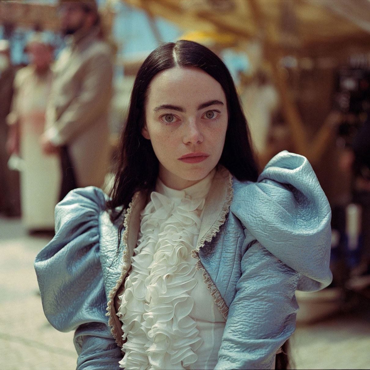 A movie still from 'Poor Things' of the actress Emma Stone. The has long brown hair, and is wearing a blue coat with large shoulders and a write top with ruffles on the front. She is looking straight at the camera.