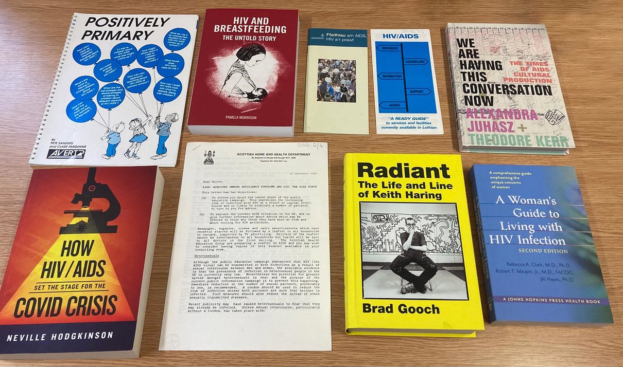 Collection of books including 'Positively Primary' by Neville Hodgkinson, 'HIV and Breastfeeding: The Unresolved Story,' 'HIV/AIDS: Are You at Risk?' by Robert J. Marzano, 'We Are Having This Conversation Now' by Alexandra Juhasz and Theodore Kerr, 'How HIV/AIDS Set the Stage for the COVID Crisis' by Neville Hodgkinson, 'Radiant: The Life and Line of Keith Haring' by Brad Gooch, and 'A Woman's Guide to Living with HIV Infection.' A central document appears to be an official letter or report.