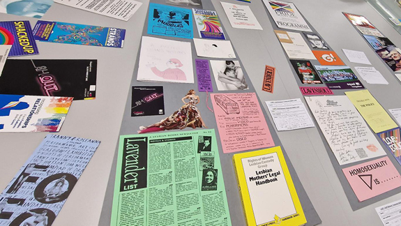 LGBTQ+ ephemera spread out on a table with people standing around it pointing at things.