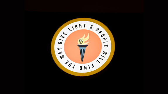 A flame framed by a circle with the words 'Give light and people will find the way'.