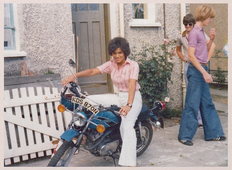 Vintage photograph of Jackie Kay sitting on a motorbike outside a pebble dashed house, smiling. She's wearing a pinkish shirt and white bell bottoms.