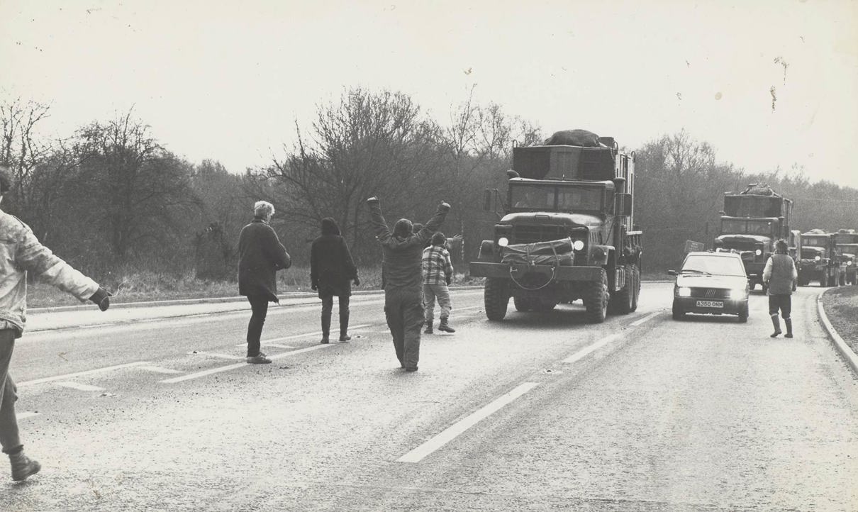 A group of people standing on a road, intercepting a convoy of large trucks. Some individuals have their arms raised.